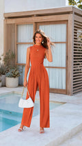 A woman stands by a pool wearing a sleeveless rust-orange jumpsuit with a high neckline and wide legs. She is holding a white handbag with a gold chain strap and wearing clear high-heeled sandals-blackorvani