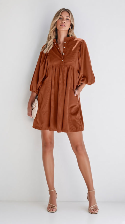 A woman modeling a rust-brown short dress with balloon sleeves and a buttoned neckline, standing against a plain light gray studio background-blackorvani