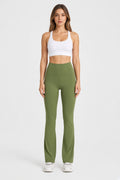 A woman stands against a light gray studio background wearing a white sports bra and olive green high-waisted flared leggings with white sneakers.