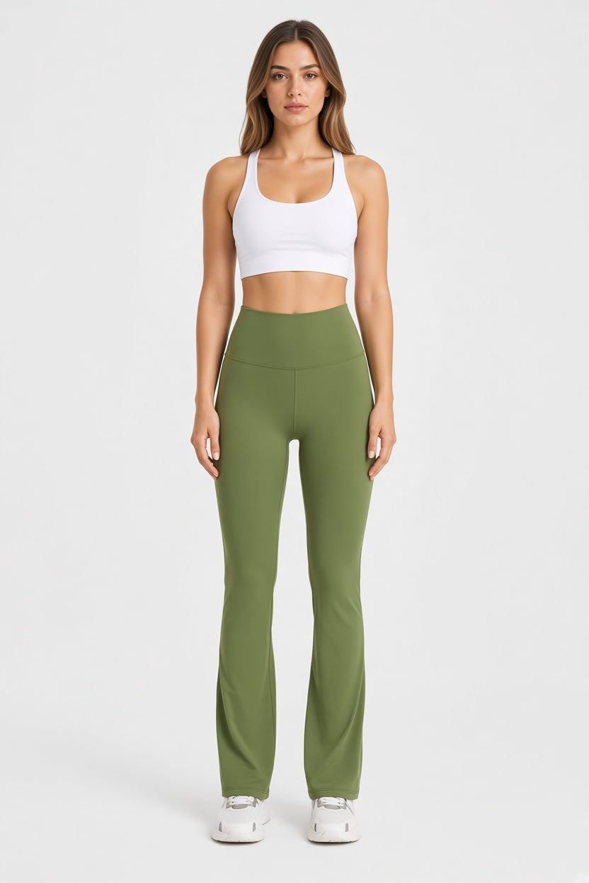 A woman stands against a light gray studio background wearing a white sports bra and olive green high-waisted flared leggings with white sneakers.